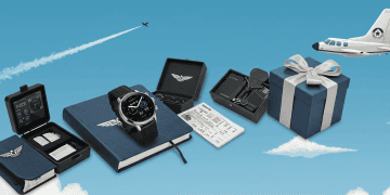 Aviation-themed gifts display against a blue sky backdrop. Features a smartwatch, notebook, sunglasses, accessories set, and a gift box with an airplane in the background.